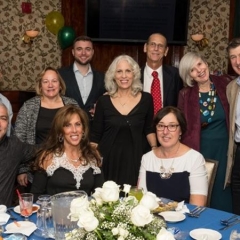 Randy Bolton, City Access Compliance Officer, poses with a group.  ID: Randy is wearing a black dress and is in the center of the picture.