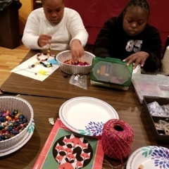 Two participants at an art class.  ID: A participant on the left is working on a necklace while the other one is looking inside a box with art supplies.