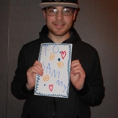 A participant holding up his artwork.  ID: A drawing with hearts that reads "To Sammy".