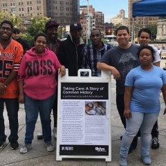 Participants pose with the sign for the exhibit.  ID: The top of the sign says "Taking Care: A Story of Common History".
