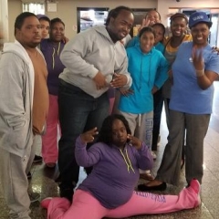 Participants wearing athletic wear pose for a group picture.  ID: A girl in the front is making a split and is holding up her fingers to form a peace sign.