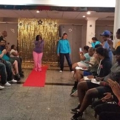 A participant strikes a pose on the red carpet.  ID: The rest of the participants are sitting to the sides of the carpet and are clapping.