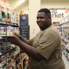 A participant smiles as he is stacking shelves at Petco.  ID: A man stands in an isle of Petco.
