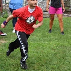 A participant is running through a ballpark.  ID: A participant is wearing a red shirt with a picture of a soccer ball on it.