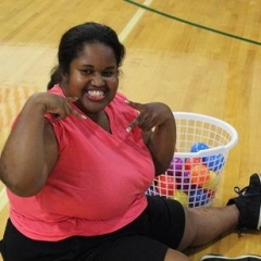 A participant smiling for the camera.  ID: The participant is wearing bright pink shirt and is sitting on a basketball court with a basket of colorful balls next to her.