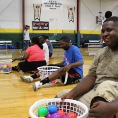 Participants with baskets full of colorful balls.  ID: Participants sitting on a floor of a basketball court.