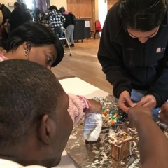 Three participants are decorating ginger bread houses.  ID: Participants are leaning over a table.
