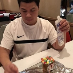 Participant decorating a ginger bread house.  ID: Participant is wearing a white shirt and is holding a tube with frosting.