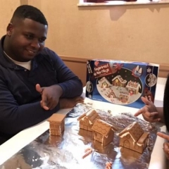 Participant smiles and points at ginger bread houses he has helped make.  ID:  The participant is wearing a dark sweatshirt.