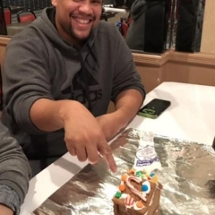 Participant is pointing to a ginger bread house he has helped make.  ID: Participant is wearing a gray sweatshirt.