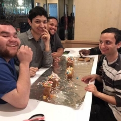 Participants smile for the camera.  ID:  Participants sit around a table with ginger bread houses that they have made.