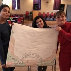 Participants hold up a table cloth that they made themselves.  ID:  The cloth reads "Home Sweet Home".