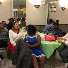 Participants sit at a table.  ID: A participant in the center is wearing a blue cheerleader costume. The one on the right is wearing bunny ears.