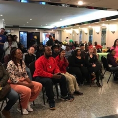 Group of participants listen to a presentation.  ID: Participants sit in chairs while listening.