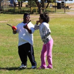 Participants playing with a nerf football.  ID: Two participants standing in a field while one of them hold two nerf footballs.