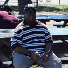 A participant enjoying a quite moment in the shade.  ID: A participant wearing a striped shirt is sitting on a bench.