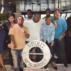 A group of participants smile for a group picture.  ID: Participants stand on board of a cruise in front of a life saver that reads "Spirit New Jersey 2017".