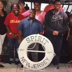 A group of participants smile for a group picture.  ID: Participants stand on board of a cruise in front of a life saver that reads "Spirit New Jersey 2017".