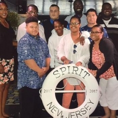 A group of participants smile for a group picture.  ID: Participants stand on board of a cruise in front of a life saver that reads "Spirit New Jersey 2017".