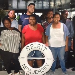 A group of participants smile for a group picture.  ID: Participants stand on board of a cruise in front of a life saver that reads "Spirit New Jersey 2017".