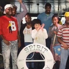 A group of participants smile for a group picture.  ID: Participants stand on board of a cruise in front of a life saver that reads "Spirit New Jersey 2017".