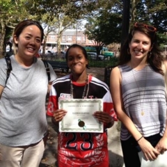 Day Habilitation participant holds up an award she received for participating in Therapeutic Garden Project for People of All Abilities.  ID: The participant stands in the middle and smiles for the camera.