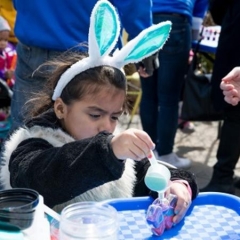 A little girl at the crafts table.  ID:  The girl is wearing blue bunny ears.