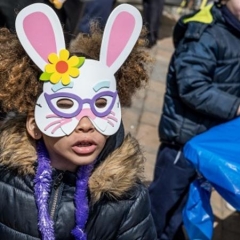 A little girl is wearing an elaborate bunny mask.  ID: The bunny mask has pink ears, lavendar glasses and a yellow flower on it.