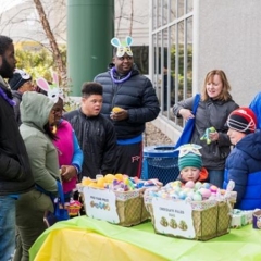 Kids wait for the Egg Hunt to begin.  ID: Egg Hunt participants stand by a table with Easter Egg baskets on it.