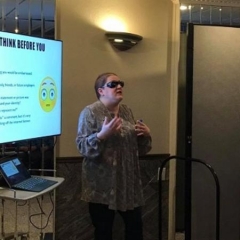 ID: Holly Bonner stands in front of a tv PowerPoint presentation that reads, "Always think before you share."