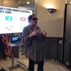 ID: Holly Bonner speaking to participants with a TV PowerPoint behind her highlighting social media platforms.