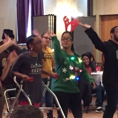 Participants perform a dance during the holiday party celebration.  ID: Participant is wearing a green sweater with colorful ornaments on it.