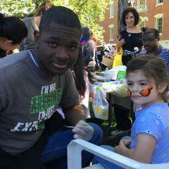 A little girl in a blue shirt is getting her face painted  at the Staten Island Snug Harbor Cultural Center & Botanical Gardens.
