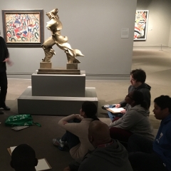 Participants listen to their teacher.  ID: Sitting in a semi-circle attentively listening during an art class.
