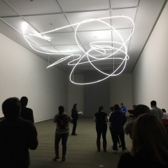 Participants roam around an exhibition room.  ID: There is a squiggly light fixture hanging from the ceiling.