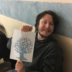 Participant smiles for the camera.  ID: He is holding up his family tree that was painted earlier in an art class.