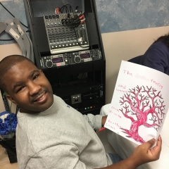 Participant smiles for the camera.  ID: He is holding up his family tree that was painted earlier in an art class.