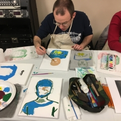 Participant painting a self portrait.  ID: Participant wearing glasses sits at a table with art supplies.